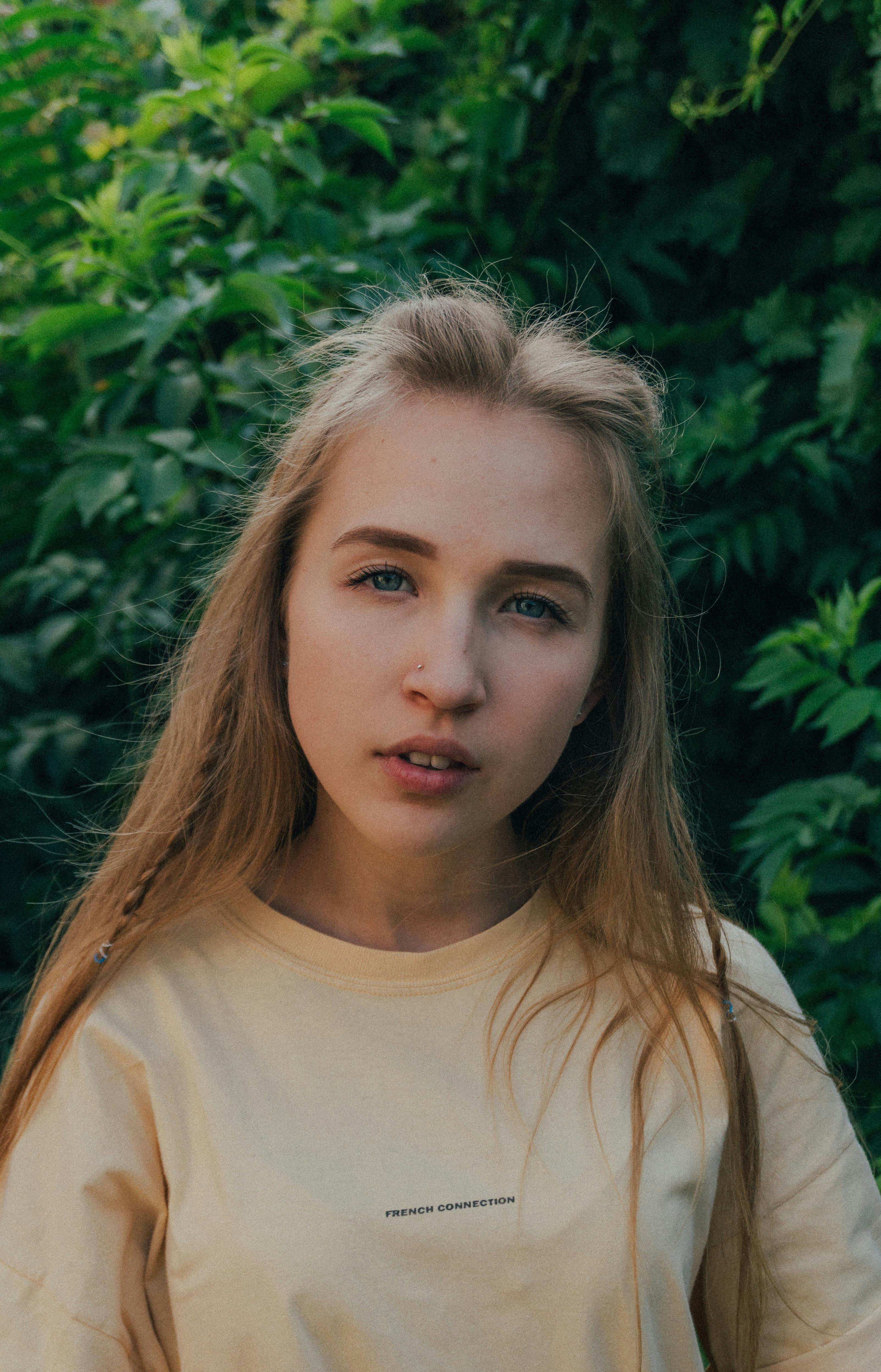 Calm female teenager without makeup with long blond hair in casual outfit looking at camera against lush green plants in garden on sunny day