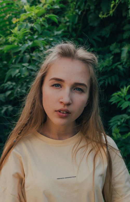 Free Calm female teenager without makeup with long blond hair in casual outfit looking at camera against lush green plants in garden on sunny day Stock Photo