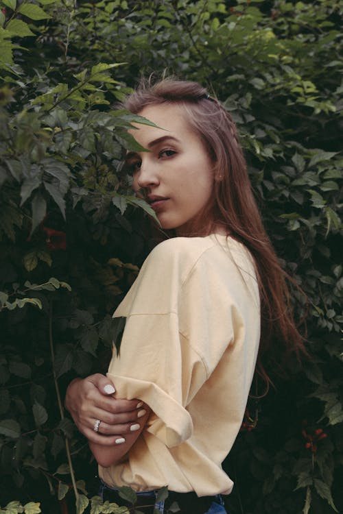 Side view of tranquil young female millennial with long brown hair in casual outfit standing near lush green shrubs with folded arms and looking at camera in garden