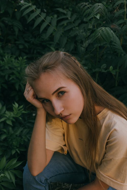 Pensive young lady sitting on haunches near lush green bushes on woods