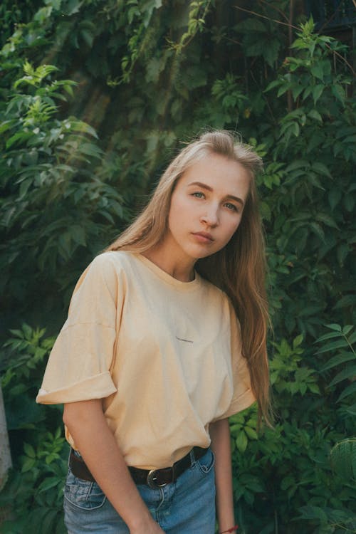 Young stylish female millennial with long blond hair in casual outfit looking at camera while spending free time in lush green garden