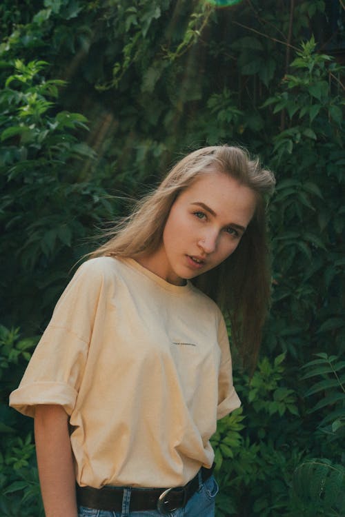 Confident young blonde woman standing near lush green foliage and looking at camera