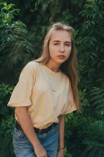 Pensive young blonde lady with long hair in trendy outfit standing near lush green trees in garden and looking at camera thoughtfully