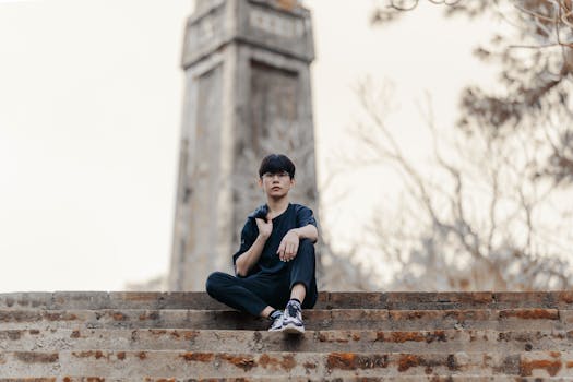 Young man sitting on stone steps with a historical tower in the background. Autumn setting, casual attire.