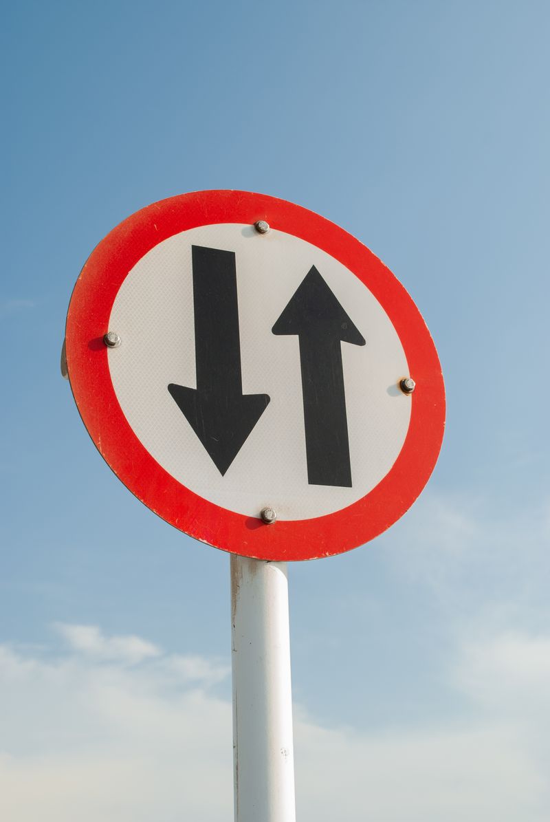 Circular two way traffic sign with arrows pointing in both directions against a clear sky