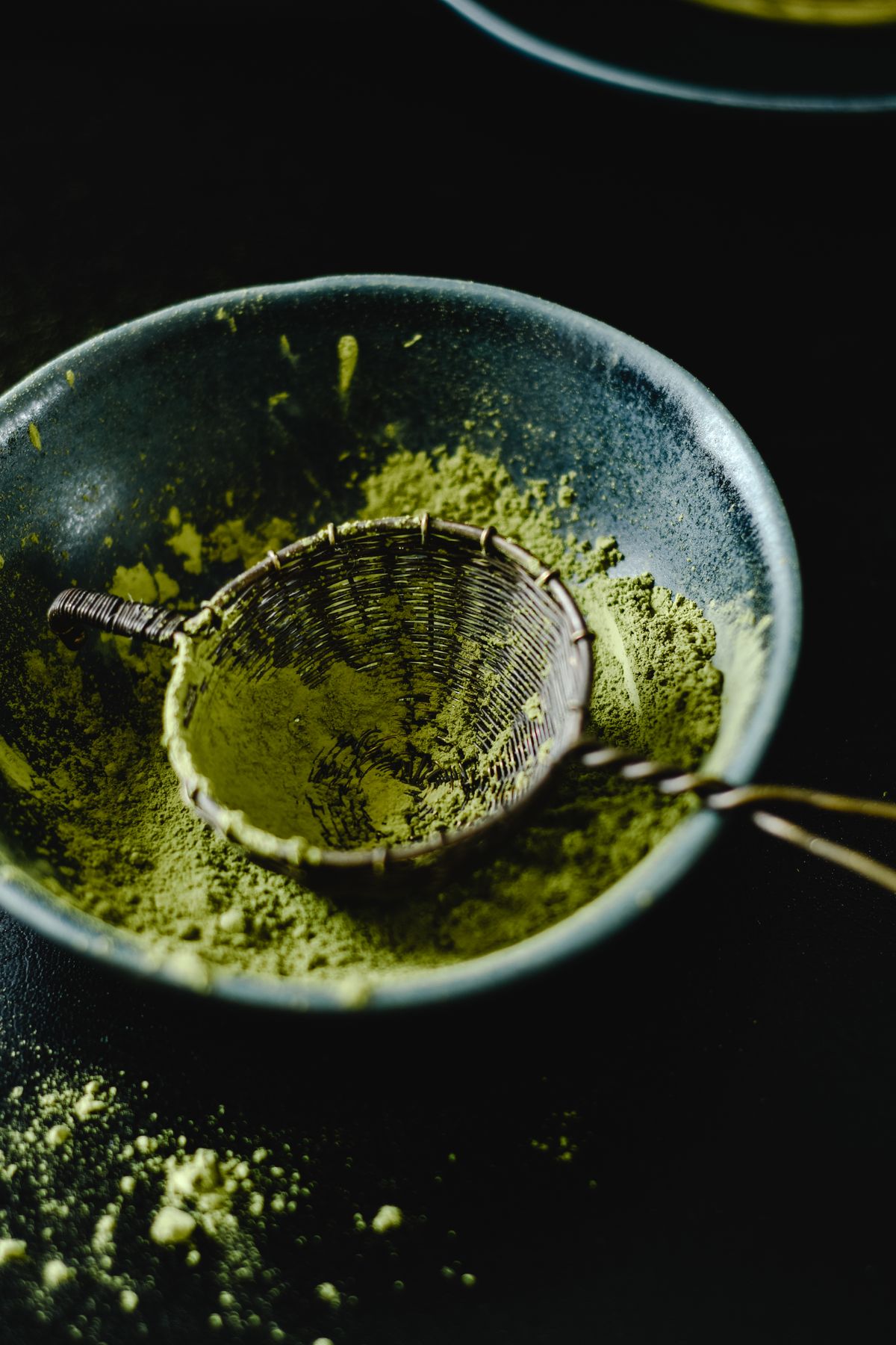 Matcha-Pulver in einer dunklen Keramikschale mit traditionellem Bambussieb – intensiv grünes Ceremonial-Grade-Pulver