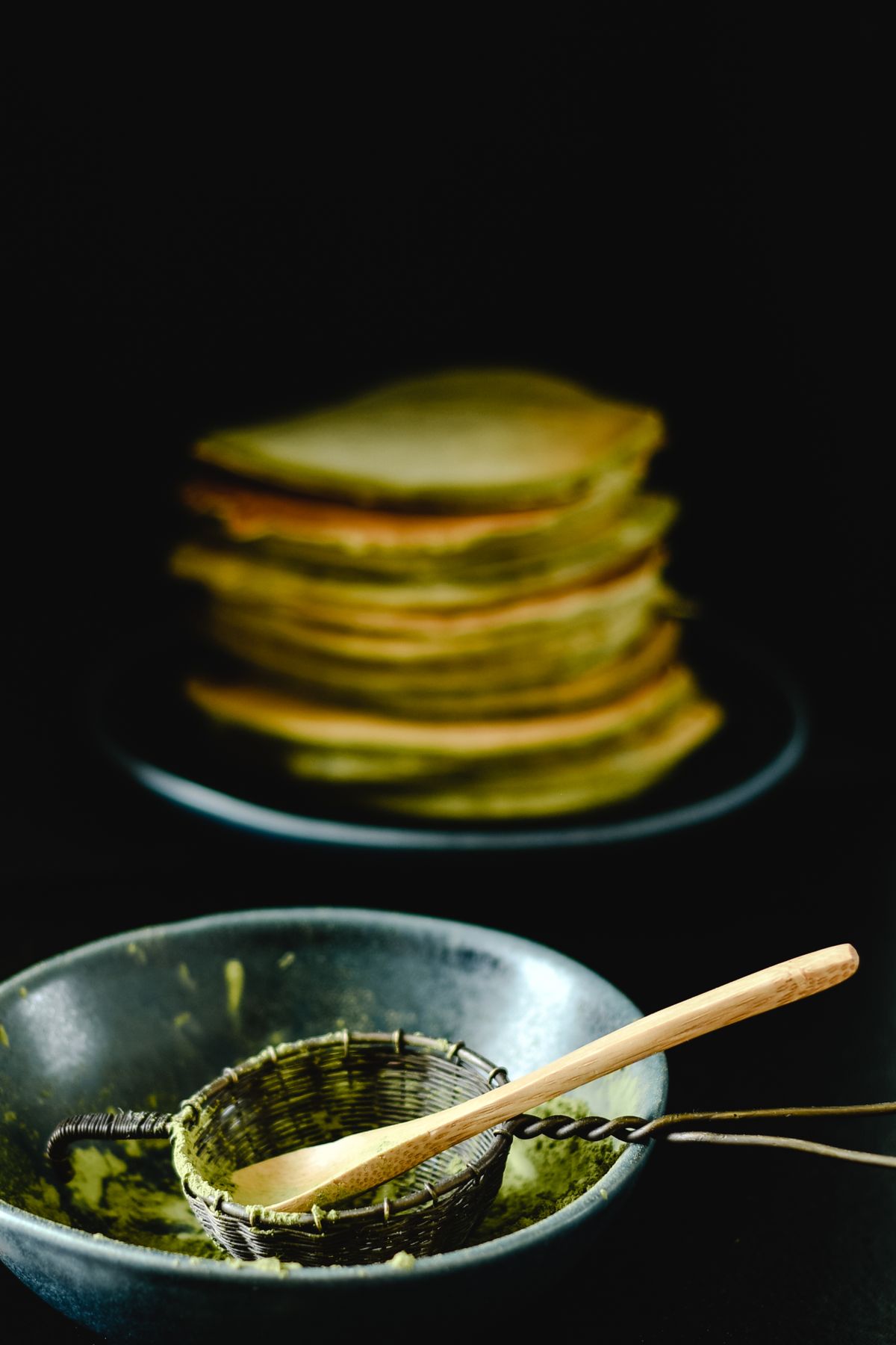 Matcha powder in a metal sifter with stacked green pancakes in the background
