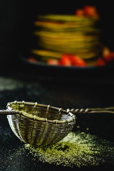 Detailed close-up of matcha powder in a metal sieve with blurred pancakes in the background.