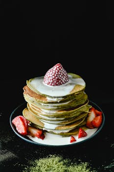 A stack of matcha pancakes topped with strawberries and cream, set against a black background.