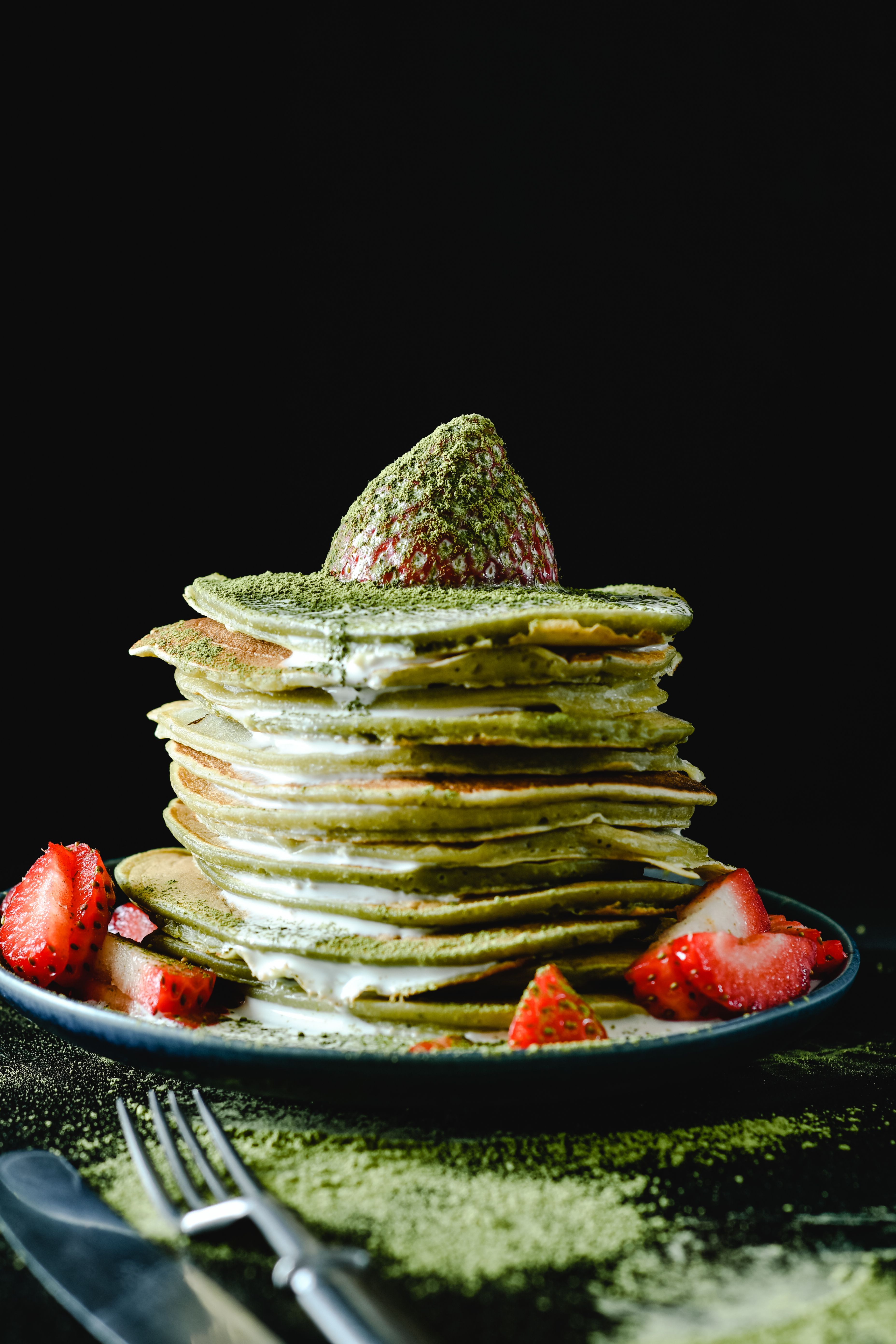 A stack of matcha pancakes topped with strawberries on a ceramic plate with cutlery.