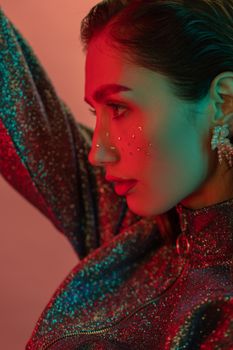Close-up profile of a woman in sparkling outfit and makeup under colorful studio lights.