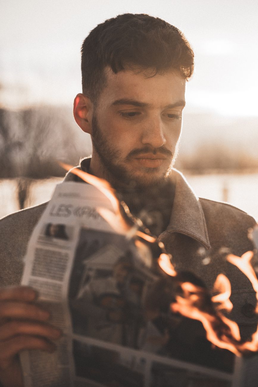 A man with a beard examines a burning newspaper in the sunlight, creating a dramatic scene.