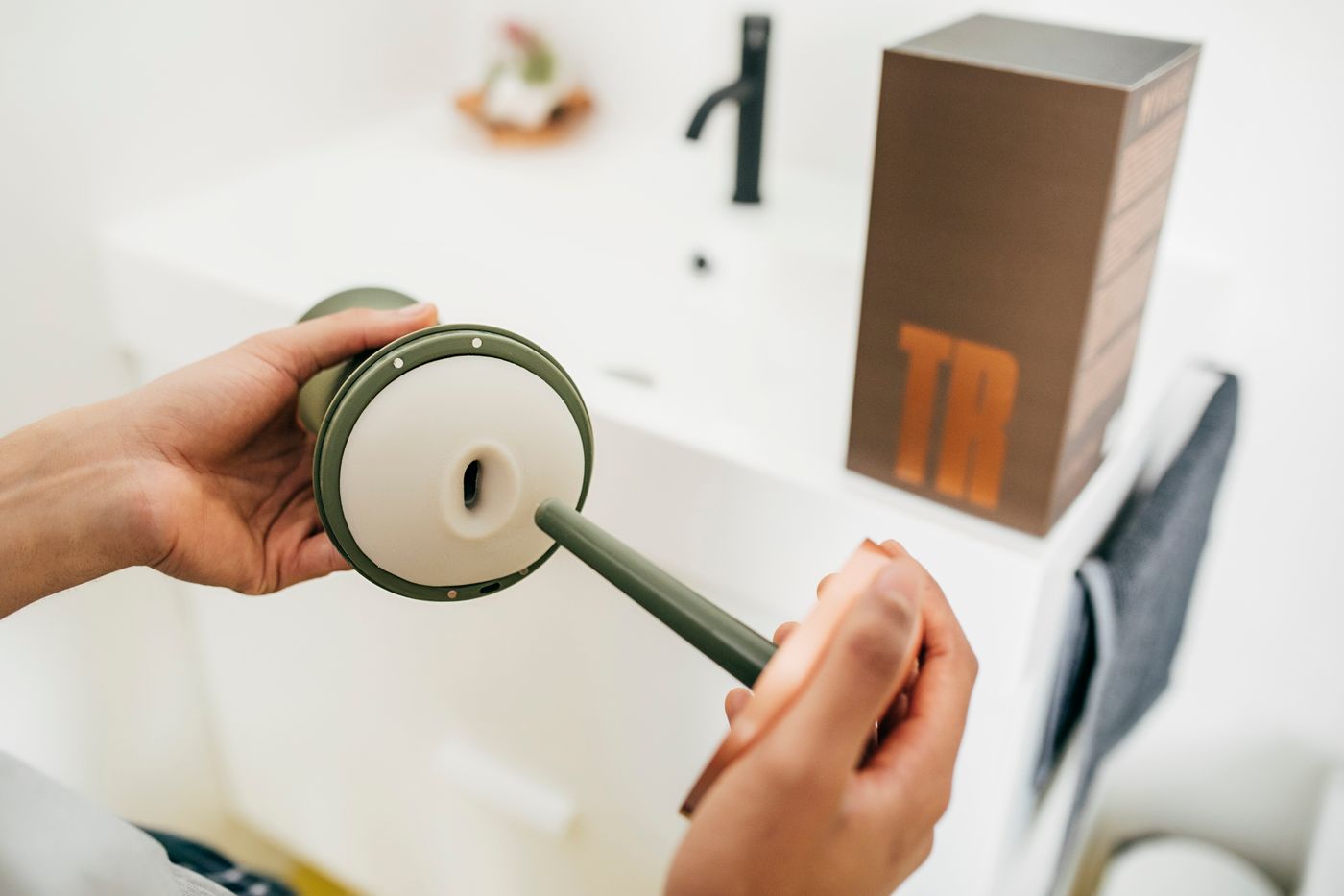 Hands assembling a green personal-care dispenser beside a clean bathroom sink