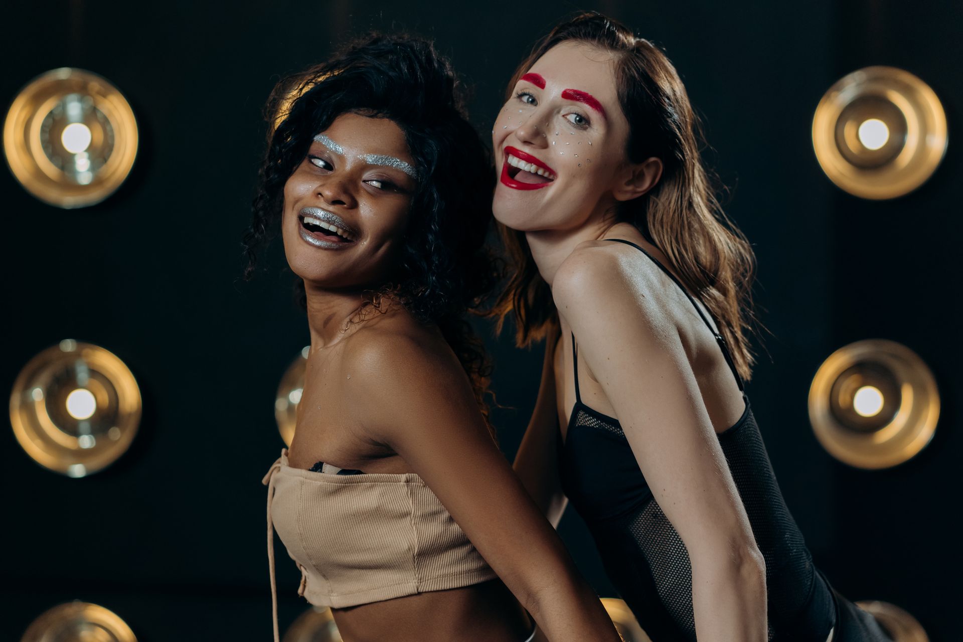 Two joyful models pose in a colorful studio setting, showcasing vibrant makeup and fashion styles.