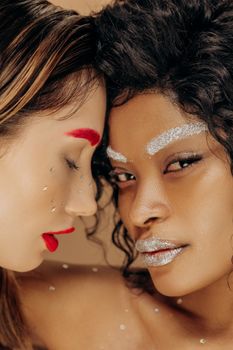 Close-up of two women with vibrant makeup and artistic glitter details in a studio setting.