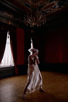 A woman in a vintage dress stands in a dimly lit ornate room, evoking a sense of mystery and elegance.