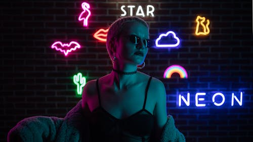 Stylish woman with sunglasses posing against neon-lit brick wall featuring modern art symbols.