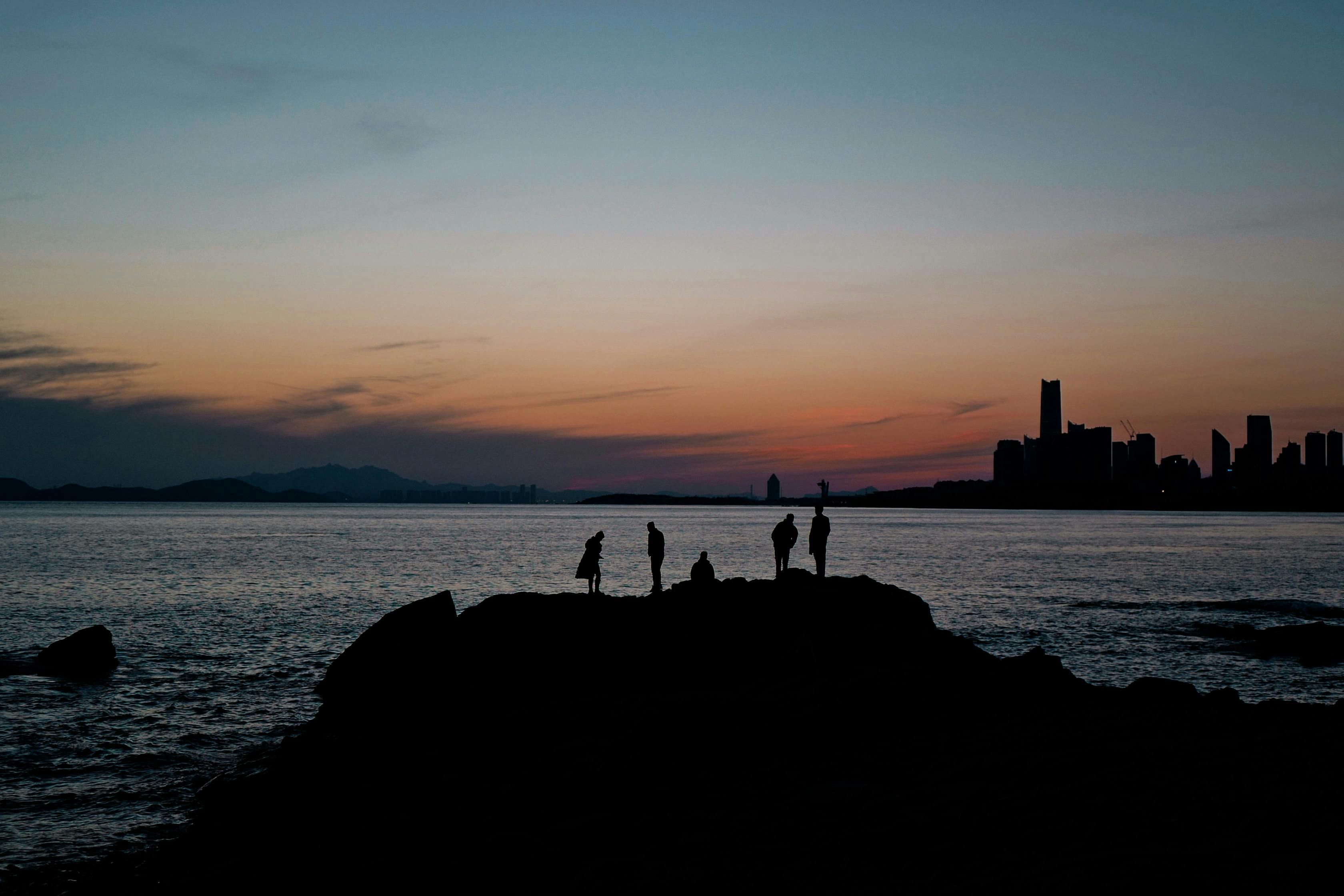 Silhouetted figures on a rocky shore during a stunning sunset with city skyline in the distance.