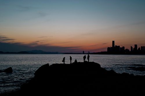 Free Silhouetted figures on a rocky shore during a stunning sunset with city skyline in the distance. Stock Photo