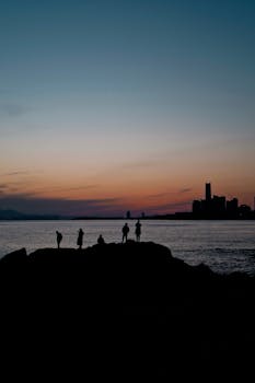 Silhouettes of people watching a tranquil sunset by the ocean shore with cityscape in the background.