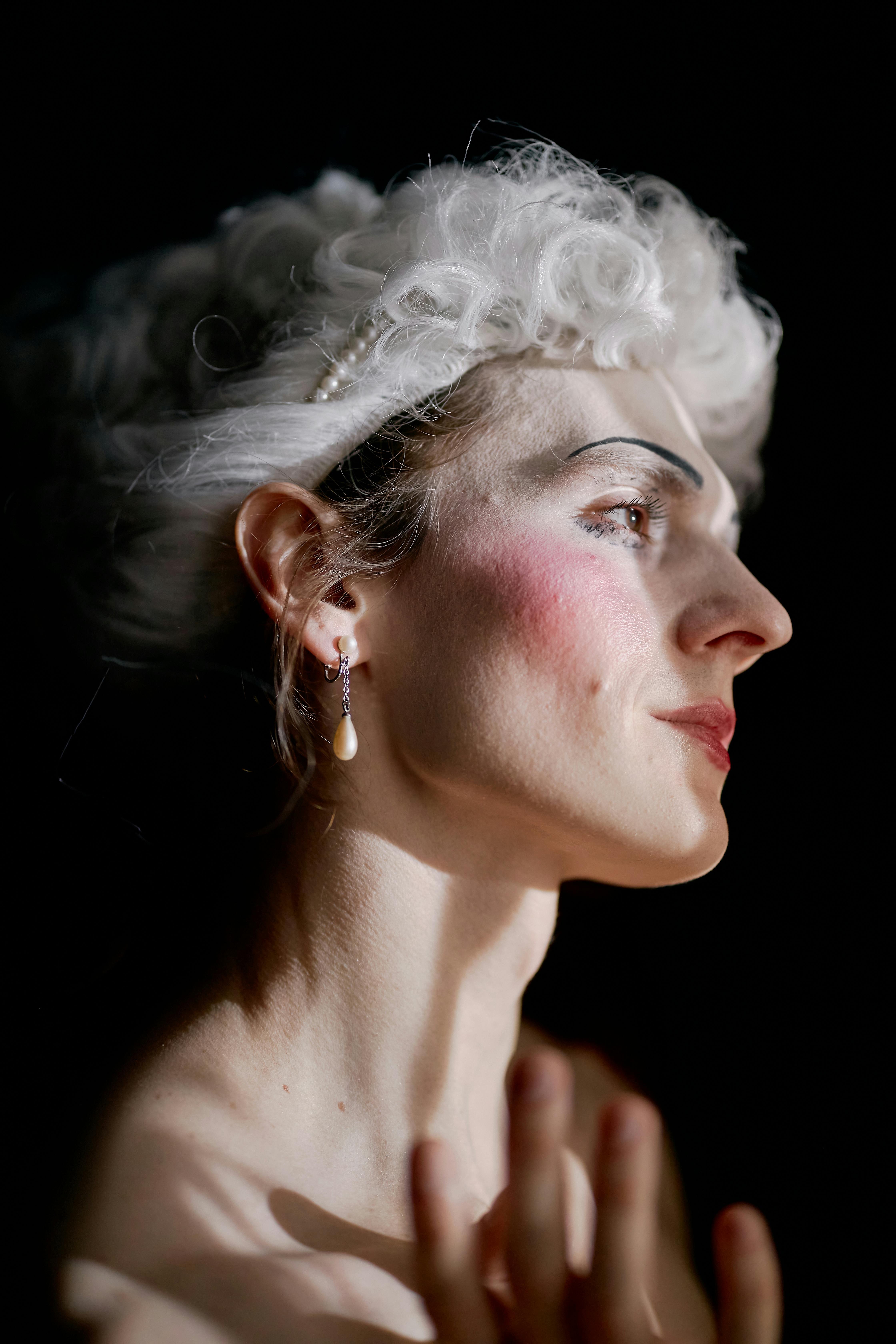 Profile portrait of a woman in theatrical makeup and costume with dramatic lighting and a dark background.
