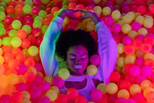 Free African American woman enjoying a colorful ball pool with neon lighting, creating a vivid and fun atmosphere. Stock Photo