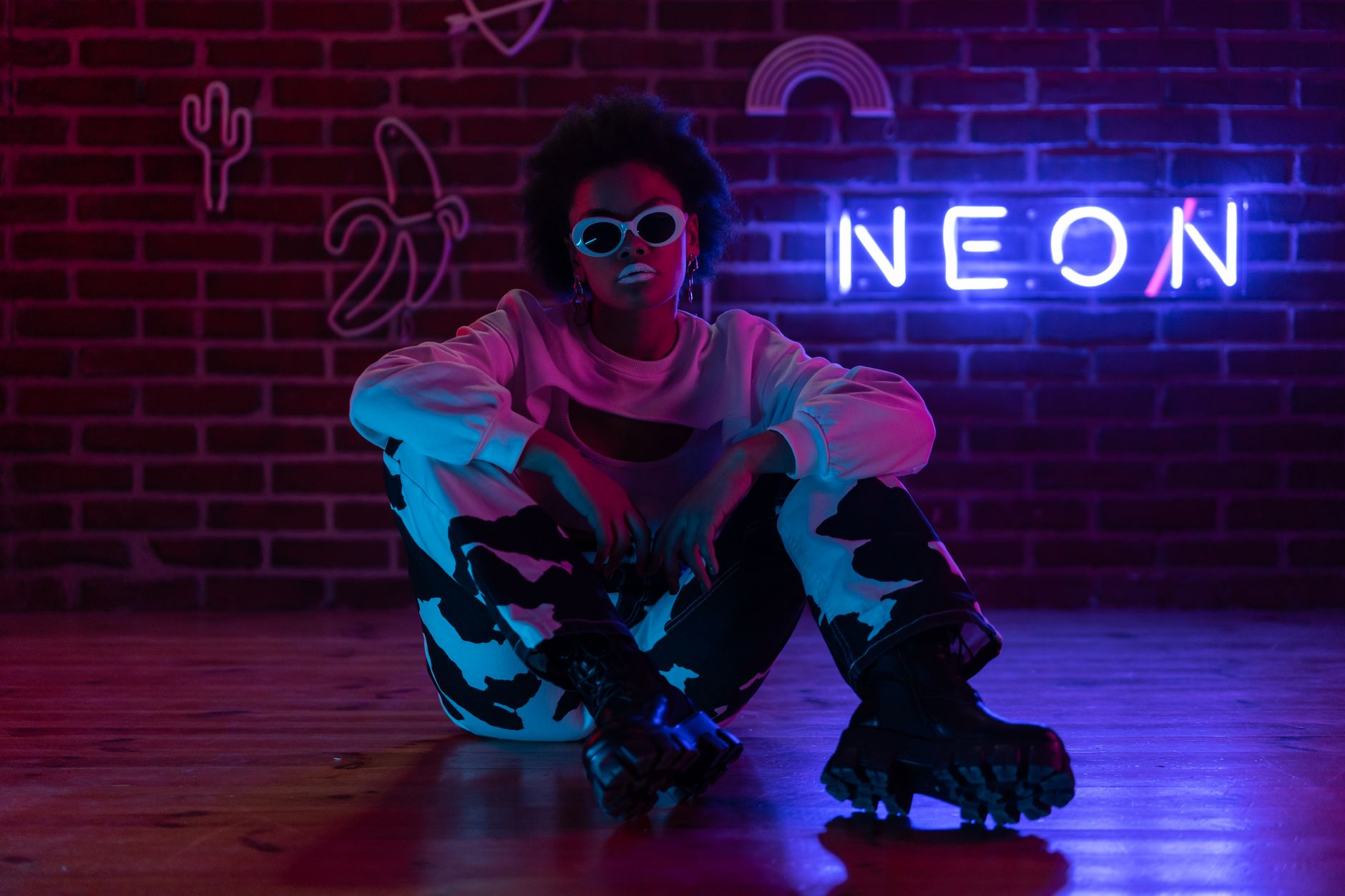 Fashionable woman with afro hair sits by neon sign, exuding style and confidence.