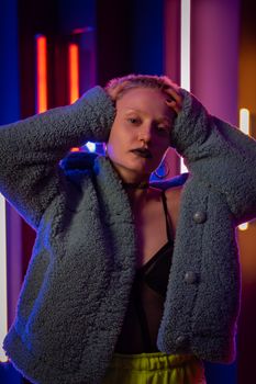 A woman poses with neon lights, wearing a textured coat and hoop earrings.