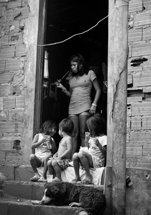 Free Black and white of young Latin American woman standing in doorway of old shabby house near little children playing on porch Stock Photo