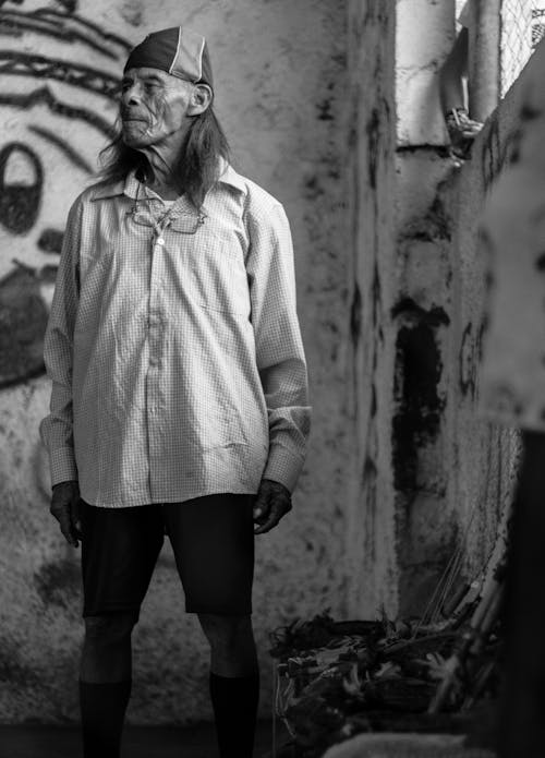 Black and white low angle of aged male standing in abandoned weathered building in poor town