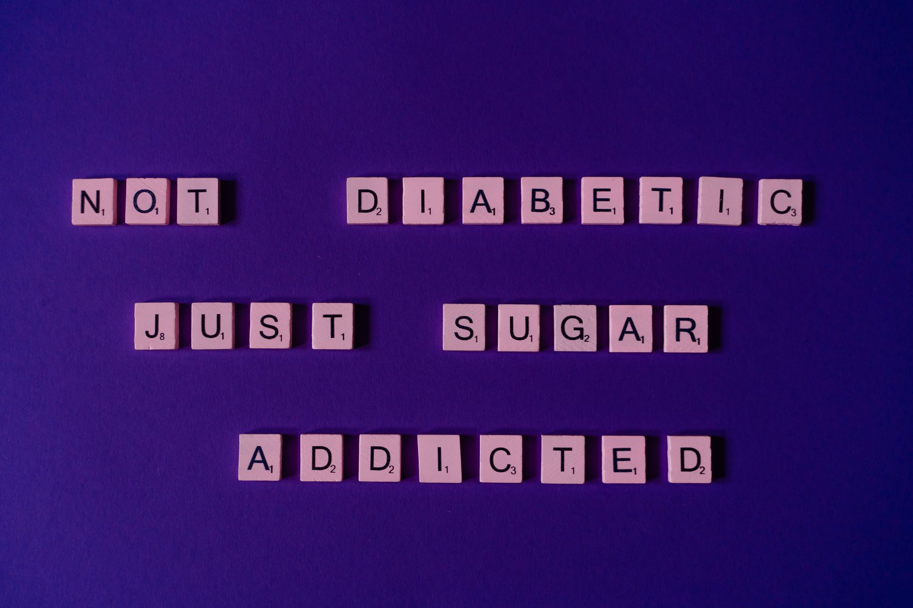 Scrabble tiles spell out a humorous message on a vivid purple background, highlighting sugar addiction.