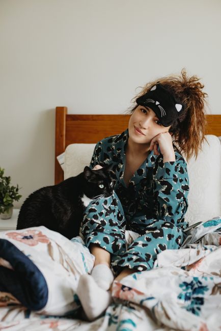 Young female in pajama and mask sitting on bed with black cat in bedroom in morning and looking at camera