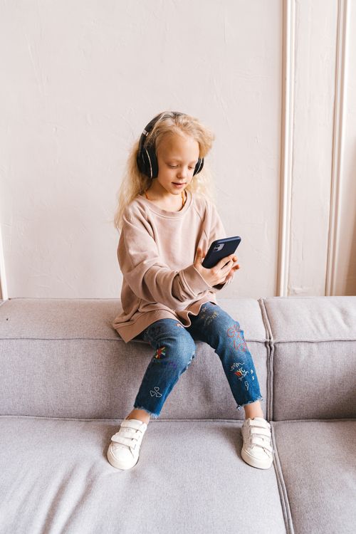 Free Blonde girl child sitting on sofa with headphones and smartphone, enjoying a relaxed indoor moment. Stock Photo