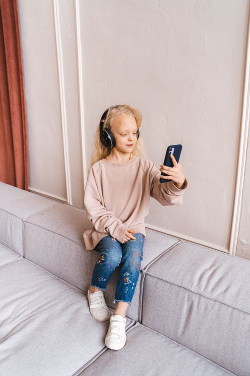 Free Child with headphones using a smartphone on a sofa, embodying tech-savvy innocence. Stock Photo