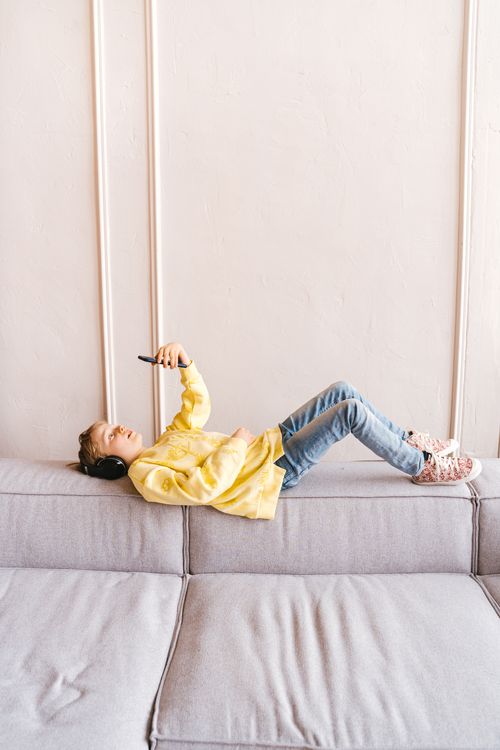 Free A young boy in a yellow sweater using a smartphone and wearing headphones, relaxing on a grey couch. Stock Photo
