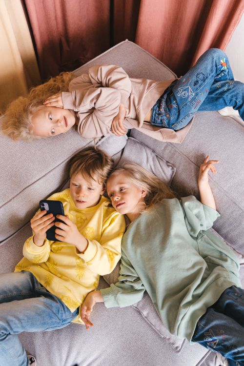 Free Three children lie on a couch, immersed in a smartphone, showcasing childhood togetherness. Stock Photo