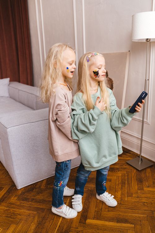 Free Two young blonde girls taking a selfie indoors with vibrant stickers on their faces, creating a playful and cute atmosphere. Stock Photo