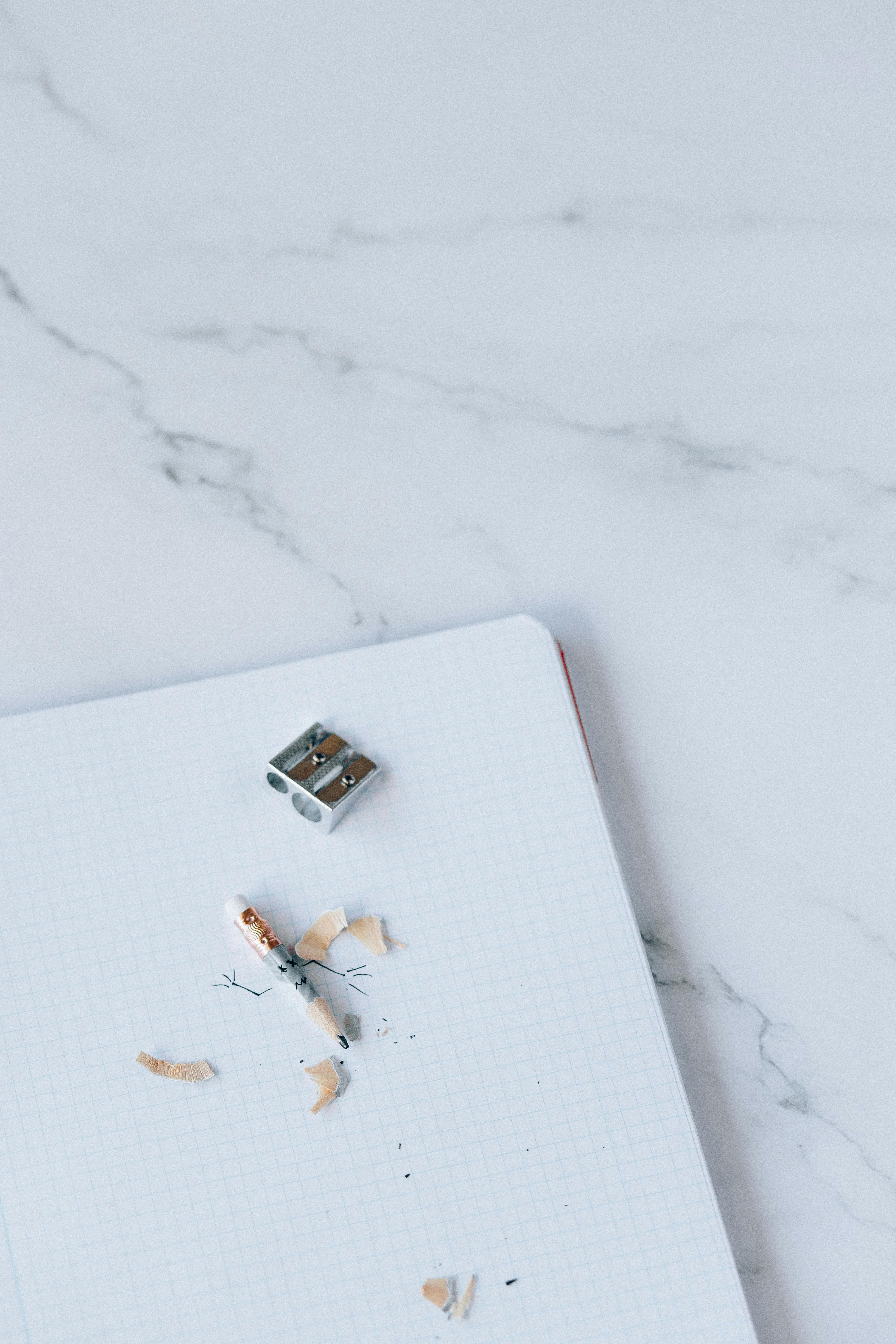 Free Top view of a notebook with pencil shavings and a sharpener on a white marble surface. Stock Photo
