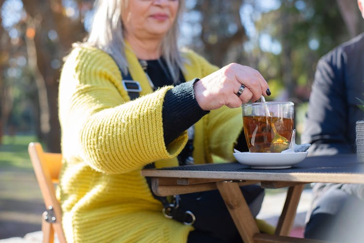 An Elderly Woman Sitting At The Park While Stirring A Cup Of Tea