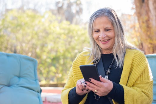 Elderly woman smiling and texting on smartphone in a sunny outdoor setting.
