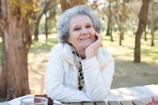 Elderly woman with gray hair smiling at a park table on a sunny day.
