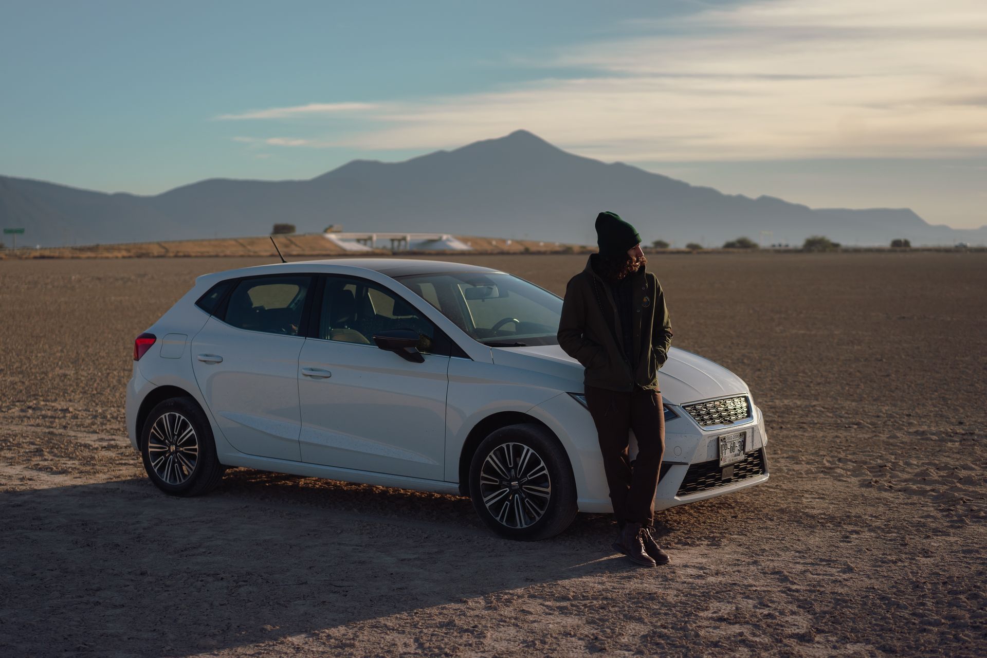 A man leans against a car in a vast desert landscape with mountains in the background.