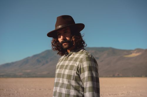 Free Man in a fedora and checkered shirt stands amidst a vast desert landscape under clear skies. Stock Photo