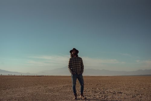 Free Man in casual attire and fedora stands in a vast desert under a clear sky. Stock Photo