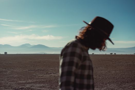 Silhouetted man in a plaid shirt and hat walking across a barren landscape with mountains in the distance.