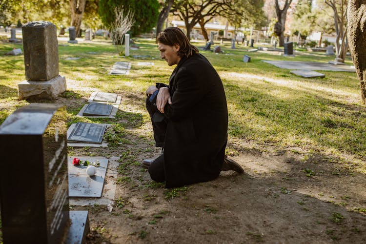 A Man In Black Coat Kneeling On A Gravestone With Ball And A Stem Of Red Rose