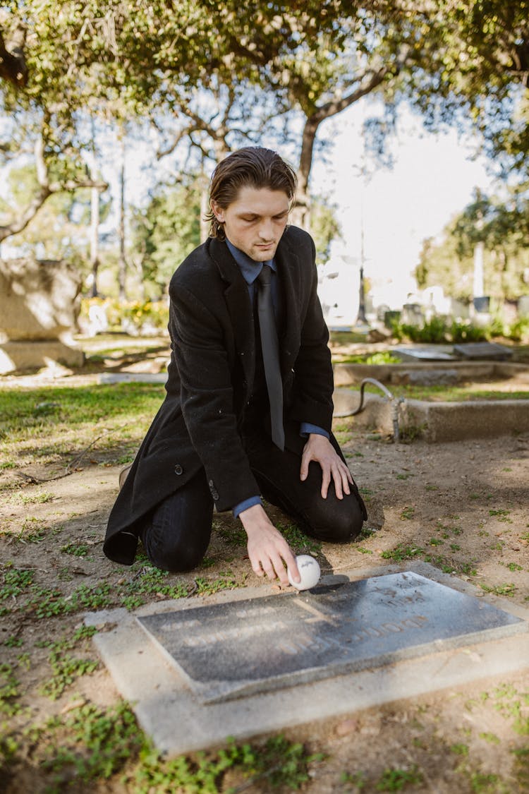 A Sad Man In Black Coat Putting A Ball On The Gravestone