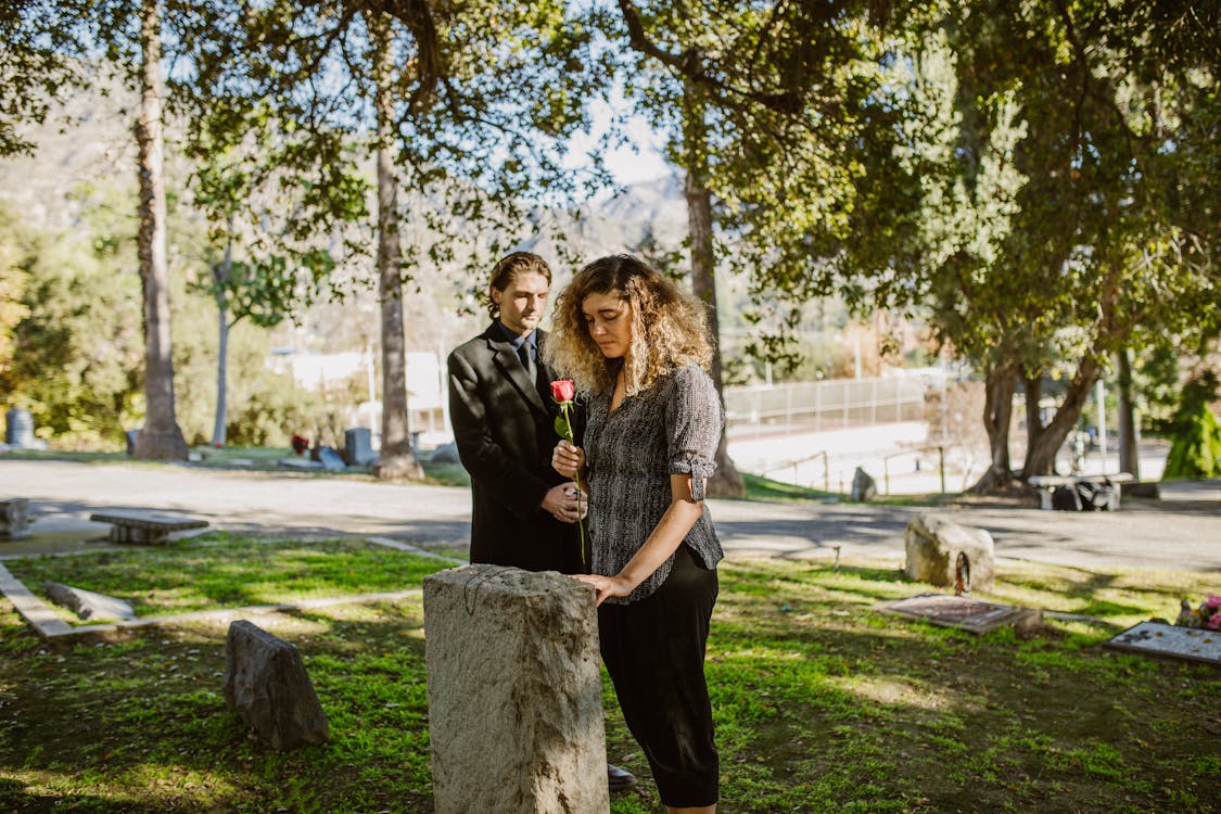  Woman in Black Shirt Standing beside Gray Concrete Tombstone while Holding Flower 