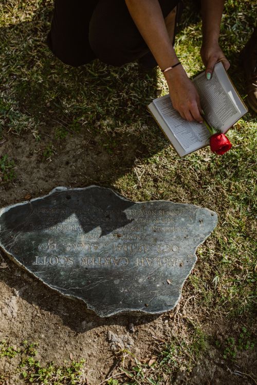 Free A woman kneels at a gravestone, reading a book with a red rose, symbolizing remembrance. Stock Photo