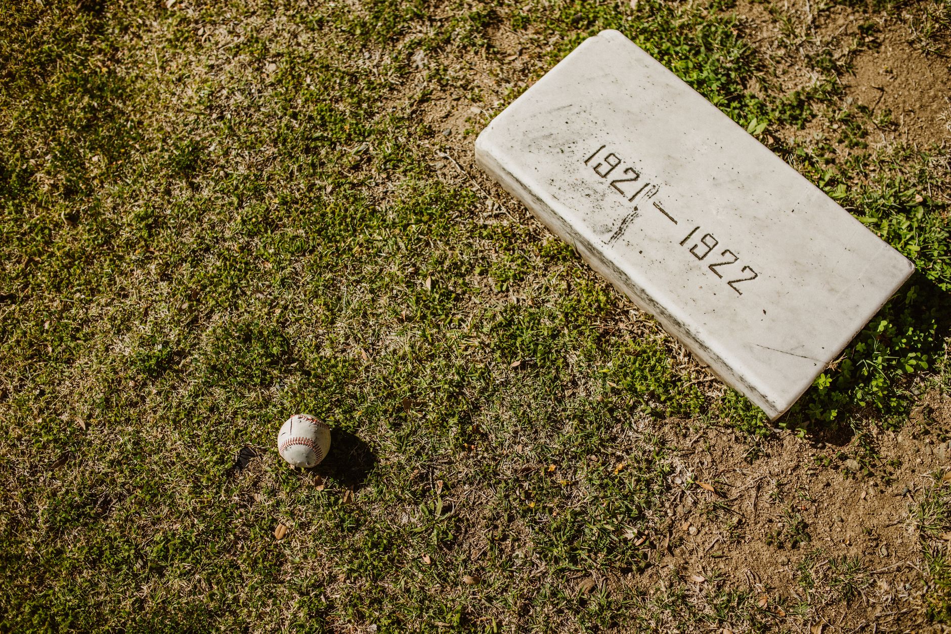 A baseball lies near an old gravestone dated 1921-1922 on a grassy field.